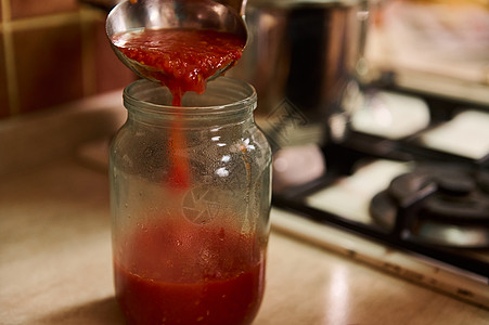 黑厨师围裙上的近身家庭主妇用一个拉盘 把锅子里煮开的番茄汁倒进消毒罐图片