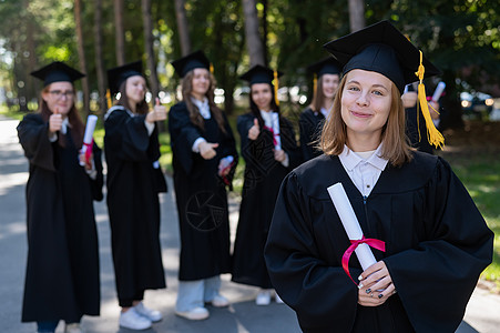 中学学生一群在户外穿毕业礼服的快乐学生 一个年轻女孩 她手上拿着文凭 在前台女性庆典公园仪式研究生证书友谊喜悦学士同学背景