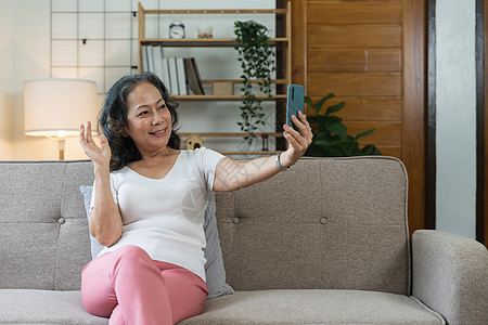 亚洲年长妇女坐在沙发上微笑着笑着坐着时在手机上自拍相机祖父乐趣女性祖父母老年退休男人幸福长老图片