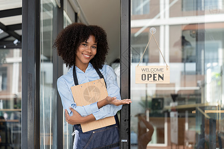 小企业非洲女老板面带微笑 同时转向咖啡馆开业的标志 穿着围裙的快乐的美国黑人女服务员企业家在门上签名工作成人职业人士服务店铺咖啡图片