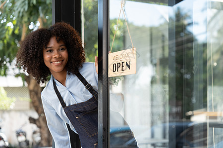 小企业非洲女老板面带微笑 同时转向咖啡馆开业的标志 穿着围裙的快乐的美国黑人女服务员企业家在门上签名入口商业员工女性餐厅咖啡师女图片