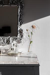 梳妆台的一部分 花瓶和花朵在白色大理石台面被阳光照亮 银色镜框镜子 带有漂亮的花押字图案 桌上放着几瓶香喷喷的香水图片