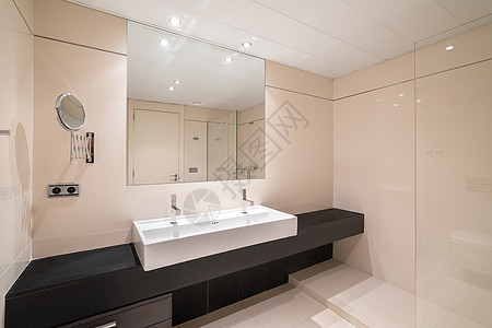 大浴室有两个陶瓷白色洗脸盆 沿着墙壁在巨大的黑色大理石台面上 墙上挂着方形镜子 镜子反射带淋浴区的入口门和带聚光灯的天花板图片