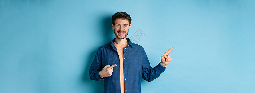 快乐笑笑的男人用左手指着徽标 展示广告 站在蓝背景上促销胡须办公室成人胡子商业工作室发型生活情绪背景图片
