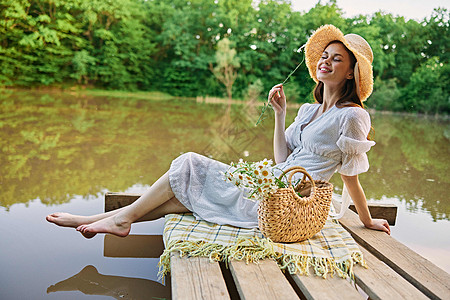 一个女人坐在湖边的码头上 穿着一件轻光衣服 戴着一顶华丽帽子 闻着香菊花味图片