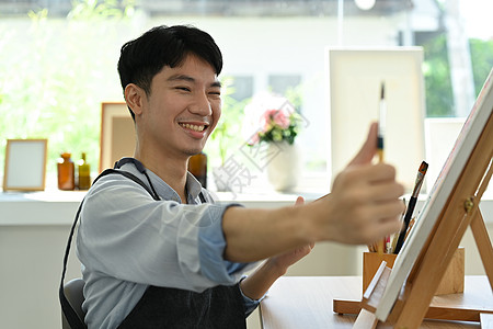 快乐的亚裔男性艺术家持有画笔和调色板绘画 张贴在艺术工作室的画布上学生画家休闲创造力活动油画阳光公寓天赋画架图片