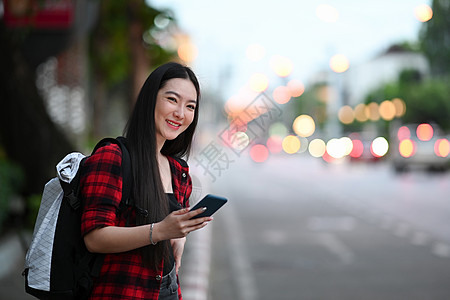 背着背包的年轻亚洲女人 用智能电话叫计程车 站在街上图片