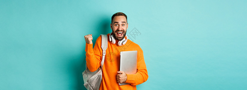背着背包和耳机的快乐男人 拿着笔记本电脑 微笑着 为胜利欢呼 战胜绿松石背景标识手势学习学生黑发潮人男性自由职业者橙子毛衣背景图片