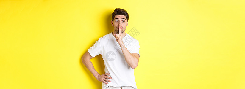 令人兴奋的男子计划出奇 对着镜头静悄悄 按手指对嘴唇用暗语标记 站在黄色背景上的照片潮人学生男性闲暇印象技术成人手势商业广告背景图片