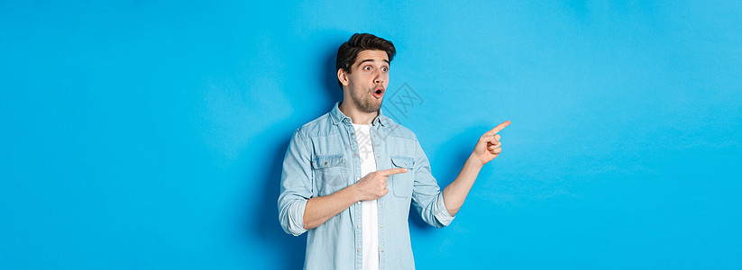 惊讶和惊奇的人在看升职 指着广告的左手指 站在蓝背景上站立横幅成功闲暇工作室情绪商业男性标识促销成人背景图片