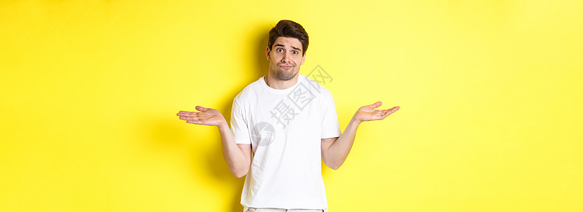 穿着白色T恤的无光男人 挥舞着迷惑不解 一无所知 站在黄色背景上黑发学生微笑男性快乐胡须技术闲暇商业工作室图片