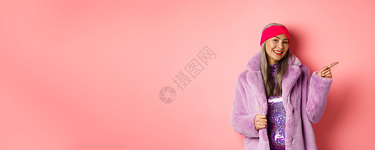 在冬毛大衣中微笑的老年妇女 看着潮流 指尖右手和展示广告 站在粉红背景下站立派对喜悦头巾购物标识毛皮女性裙子奶奶快乐图片