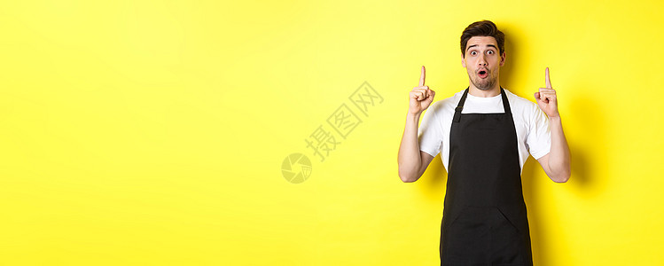 穿着黑色围裙 展示标志或广告 在复制空间指手指尖 站在黄色背景的男子服务员惊讶地看到胡须商业工作室学生快乐男性促销潮人标识闲暇友好的高清图片素材
