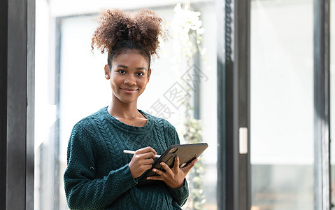 在办公室的窗户旁边站着时 却使用数码平板电脑拍摄微笑的年轻创业妇女的照片女性头发互联网爆炸屏幕商业商务人士技术工作图片
