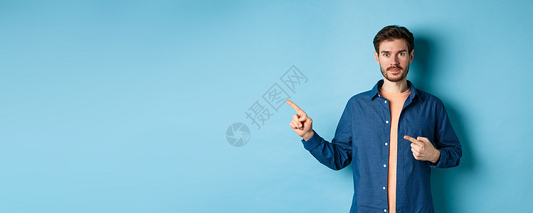 穿着衬衫的英俊年轻男子 用手指左手指着Promo 显示标志 站在蓝背景上广告快乐胡子商业男人工作室发型办公室情绪成人背景图片