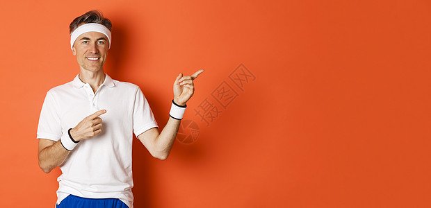 身着头带和白色 T 恤的帅气男运动员的肖像 指着右上角微笑 展示体育宣传 站在橙色背景上情感中年成人健身房橙子男性运动服训练工作图片