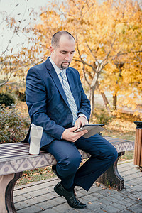 穿着西装的成熟商务人士坐在公园的长椅上看新闻 企业家在秋季公园喝咖啡 高级管理人员在城市公园休息商务自由职业者微笑电脑人士城市工图片