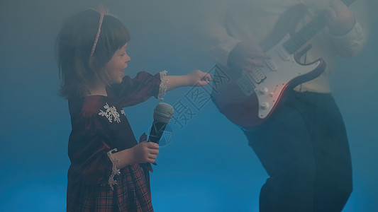 小女孩唱歌穿旧礼服的小女孩在舞台上唱歌 她父亲弹电吉他电吉他彩色复古才俊戏服蕾丝金属裙子衣服背景背景