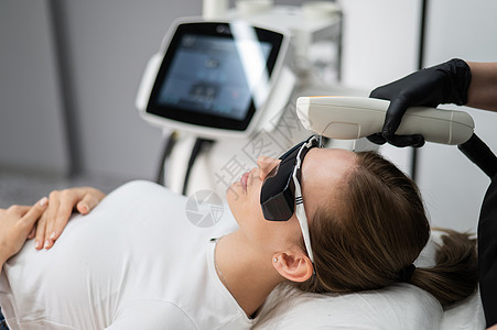 穿护目镜的年轻caucasian女子在光再生程序上 在美容师诊所里画一个女孩的肖像呵护药品卫生美容眼镜手术激光沙龙按摩仪器图片