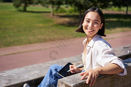 带着数字平板和图形笔坐在长椅上 笑着微笑 对着镜头转过身来并热烈说话的韩国快乐妇女公园女孩享受药片喜悦绘图自由艺术家黑发女性图片