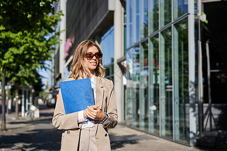 戴着墨镜和米色西装的企业女性肖像 拿着蓝色文件夹和办公室文件 去上班 在阳光明媚的日子里走在街上微笑代理人套装职场企业家员工售货图片