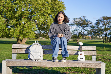 坐在公园长椅上的褐发美女 背着尤克里里琴和背包 拿着智能手机 使用移动应用音乐家女孩唱歌女性乐器弦琴音乐吉他乐趣花园图片