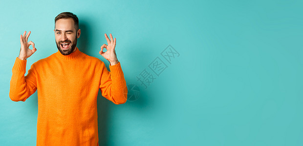 自信的男人保证一切安好 显示好的信号和对你眨眼 推荐产品 站在工作室背景的橙色毛衣购物胡须横幅广告男性橙子标识黑发胡子潮人图片