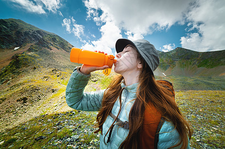 在壮丽的山景背景下 一位年轻美女留着长发 身穿夹克 头戴巴拿马帽 背着背包 用黄色瓶子喝水的头像图片