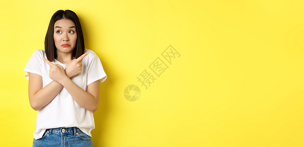 麻木不仁的亚洲女孩需要帮助选择 指指手指侧面和看不清 站在黄色背景上站立黑发女性青少年促销交易手势学生发型情感广告图片