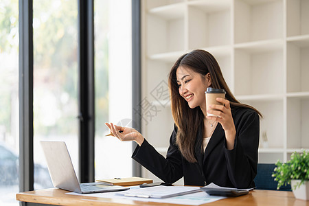 美丽的亚洲年轻女商务人士微笑着拿着咖啡杯和笔记本电脑在办公室工作 她笑着女孩杯子商业职场经理热饮技术咖啡桌子快乐图片