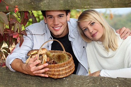 蘑菇女孩秋天在木制屏障后面的一对年轻夫妇 男子拿着一个装有蘑菇和栗子的鸡蛋篮子背景