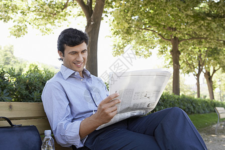 青年商务人士在公园坐在板凳上看报纸时阅读报刊图片