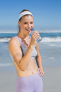 运动快乐的金发金发美女在海滩上喝水文胸护理身体假期晴天紫色女性瓶子微笑锻炼图片