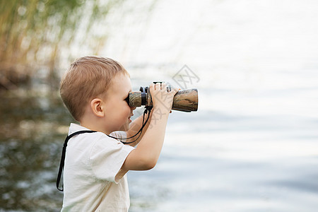 小男孩在河岸看望双筒望远镜孩子池塘间谍游客童年快乐流浪者微笑手表海岸图片