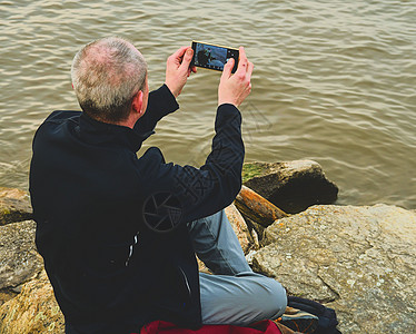 Midle 年迈男子坐在湖岸边 孤单成年男子在银行上拍自拍照片 孤独感概念蓝色微笑技术管理人员电话海岸成人经理男性城市图片