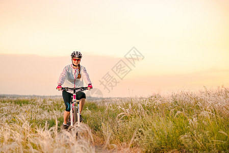 年轻女子在日落 冒险和旅行的青春草原上骑着山地自行车跑车轮羽毛娱乐山地车运动员地平线女士头盔场地天空图片