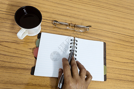 写在他的日记的商务人士 在木桌上的咖啡和眼镜旁边打开带有空白页的笔记本 商业静物概念与桌上的办公用品 带复制空间的顶视图铅笔人士图片