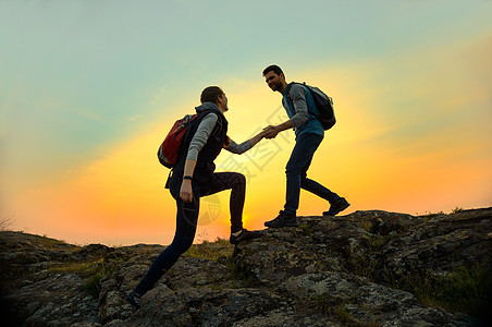 旅行者在日落山上远足 男子帮助妇女攀登顶峰 家庭旅行和冒险登山者友谊夫妻远足者悬崖风险首脑救援岩石阳光图片
