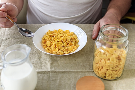 一个剪短的男人拿着一把勺子 勺子放在玉米片里 牛奶放在普通的粗糙桌布上的白盘子里 从上面看 概念 简单快速的美式健康早餐人手粮食图片