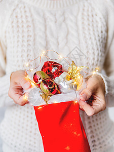 穿白色针织毛衣的女人拿着带新年装饰的红袜子 明亮的红色和金色圣诞树装饰玩具图片