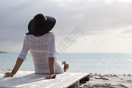 坐在海边留着长发的年轻女子在黎明时分在风中看着地平线 身着白色蕾丝连衣裙 白色内衣和大黑帽天空身体情感女性假期戏剧性孤独海景自由图片