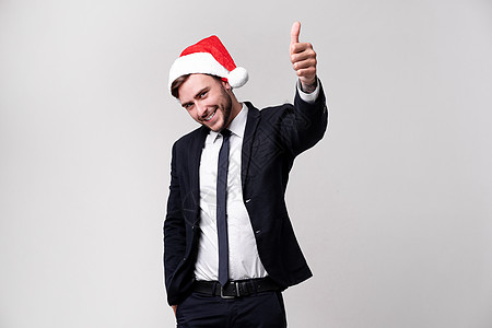 穿着西装和圣诞老人帽子的年轻英俊caucasian男子 在电影棚里站在白色背景上 微笑 露出拇指人士商业乐趣新年工作室情绪胡须情图片