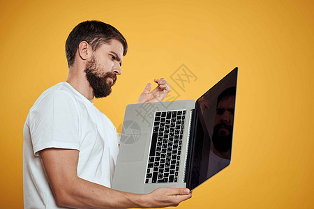 一个留胡子的男人手里拿着笔记本电脑 站在一个黄色背景键盘监控器上 监视新技术的侧观职场男性眼镜快乐幸福工作桌子商务办公室人士图片