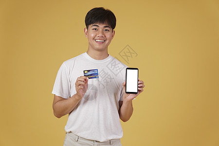 手机卡片拥有智能手机和信用卡的快乐年轻亚洲男子肖像 用黄色背景与世隔绝的网上购物卡购物者卡片消费者购物支付商业购物狂信用互联网开支背景