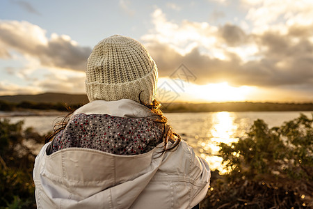 从后面看地平线的沉思人视图 从身后看到的面目全非的女人穿着冬季羊毛连帽服 看着美妙的日落或日出 海面上有云图片