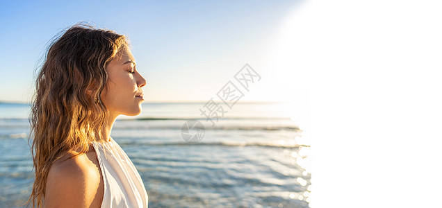 年轻美丽的白人金发女子的侧面肖像 闭着眼睛 穿着白色波西米亚风连衣裙 在日落或黎明时分在海滩上被阳光亲吻 背景是大海 白色复制空图片