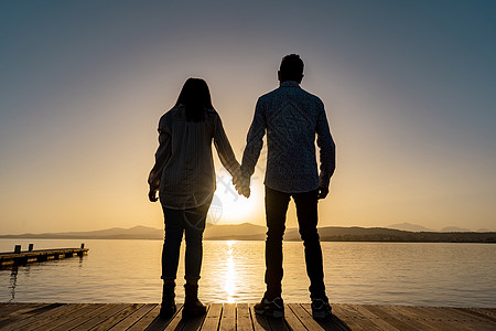 一对相爱的年轻夫妇手牵着手站在码头上 看着太阳落山或在平坦的海水中从地平线上升起的暗示性剪影背照 光线反射 正面情绪图片