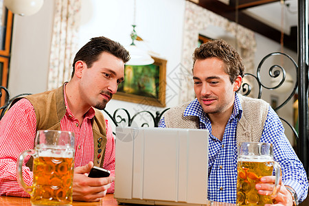 电脑和手机两个朋友在巴伐利亚酒吧 用笔记本电脑成人饮料细胞啤酒皮裤商业友谊工作餐厅传统背景