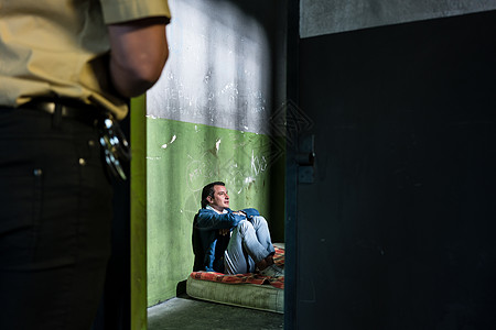 年轻男囚犯独自坐在由一名警官看守的破旧牢房里图片