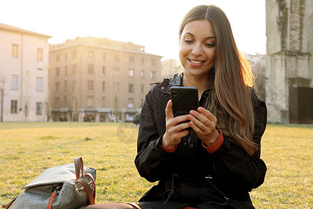 学生女孩在春季时间坐在草地短信上与手机一起户外运动电话图片